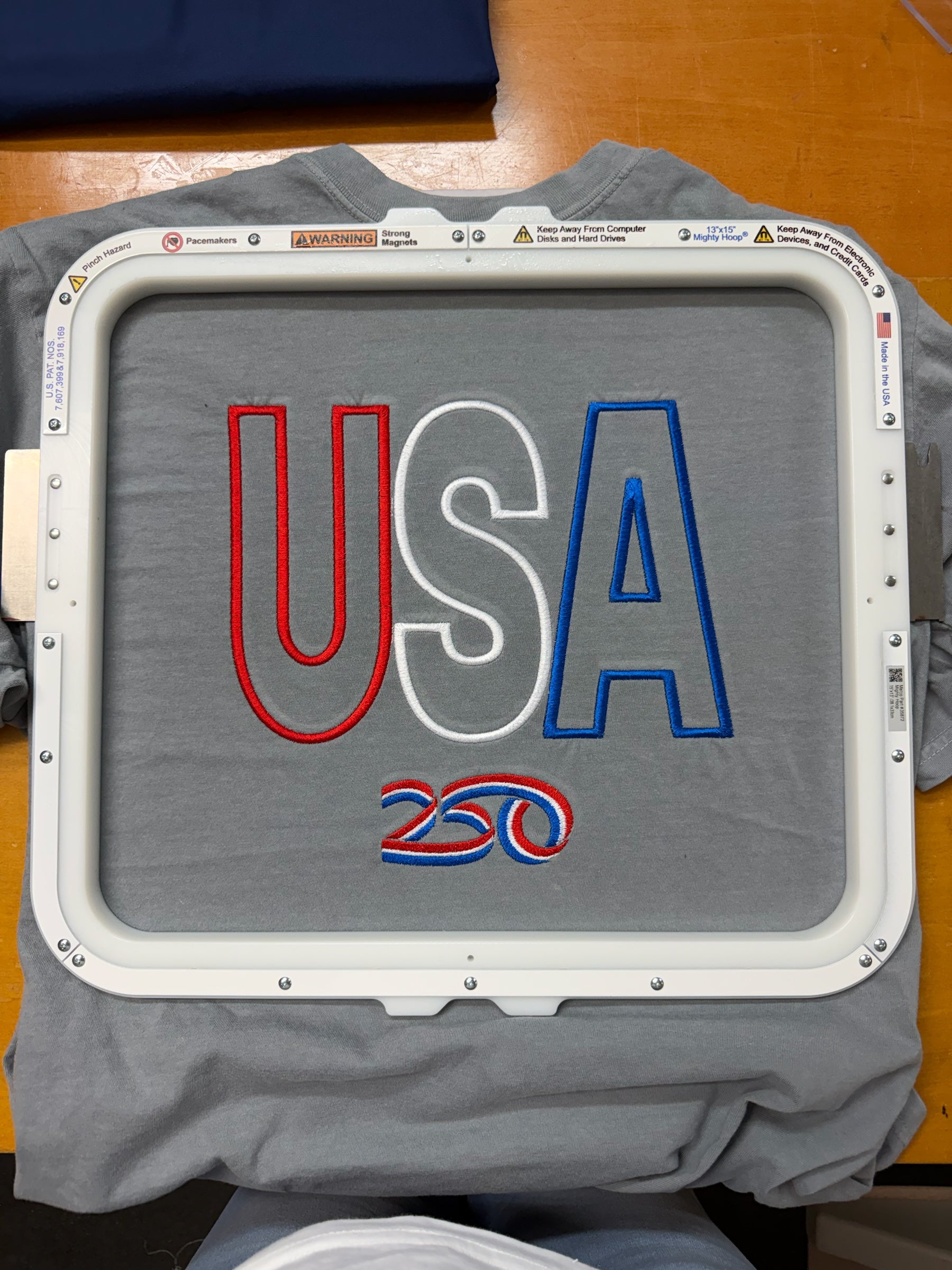 Captain Hawk Designs grey crewneck t-shirt featuring embroidered USA 250 graphic in red, white, and blue thread.